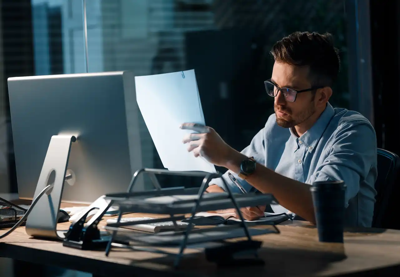 Mann mit Brille sitzt am Schreibtisch und betrachtet konzentriert ein Blatt Papier in einem abgedunkelten Büro