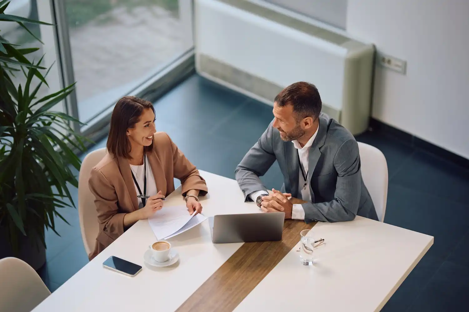 Zwei Personen in Businesskleidung sitzen an einem Tisch mit Laptop, Dokumenten, Smartphone und Kaffeetasse und unterhalten sich freundlich in einem modernen Büro.